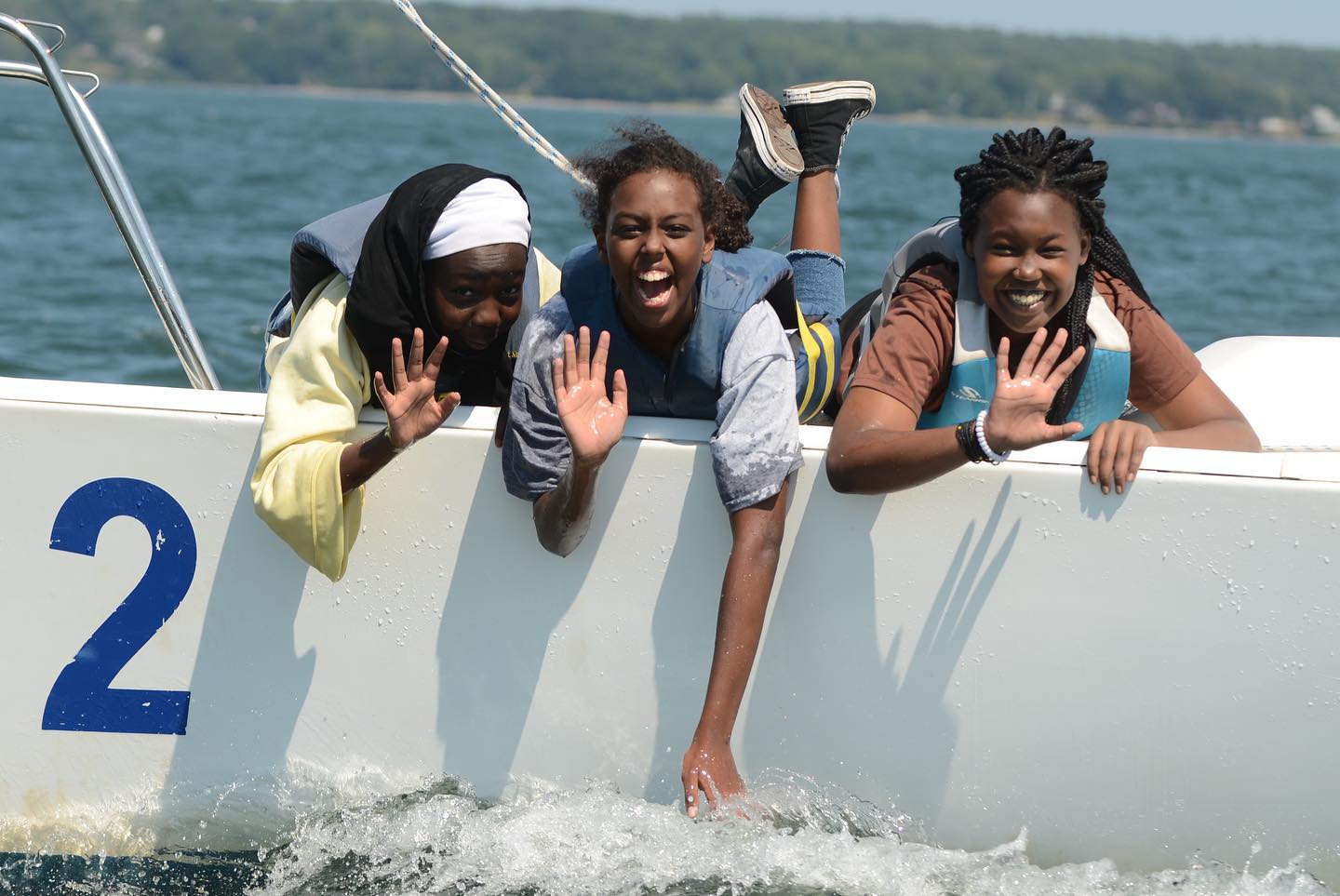 Three young people wave from the bow of a sailboat